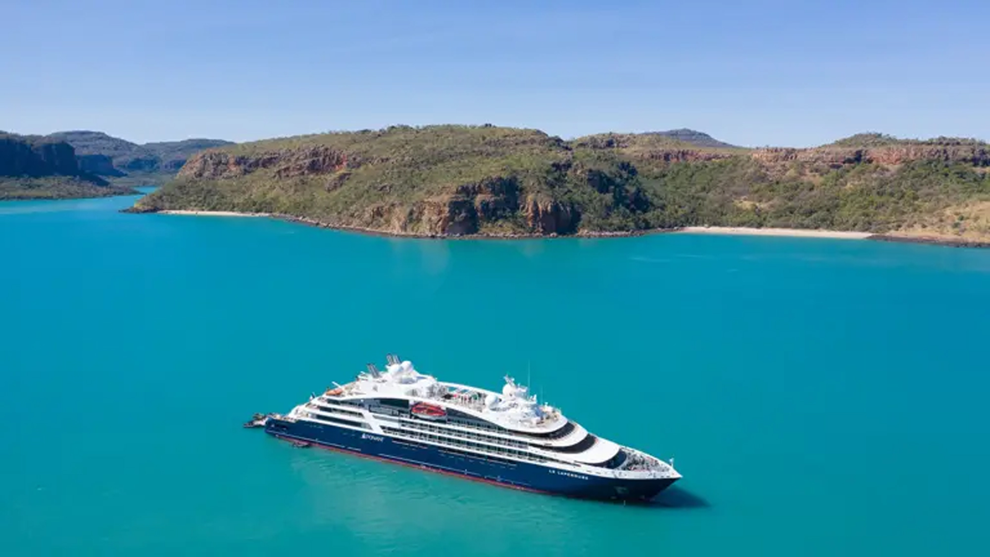 A modern expedition cruise ship anchors in pristine turquoise waters off a rugged, vegetation-covered peninsula in Australia's remote Kimberley region. The dramatic landscape features rocky outcrops, dense bushland, and a secluded white sandy beach, exemplifying the untamed natural beauty of this World Heritage area.