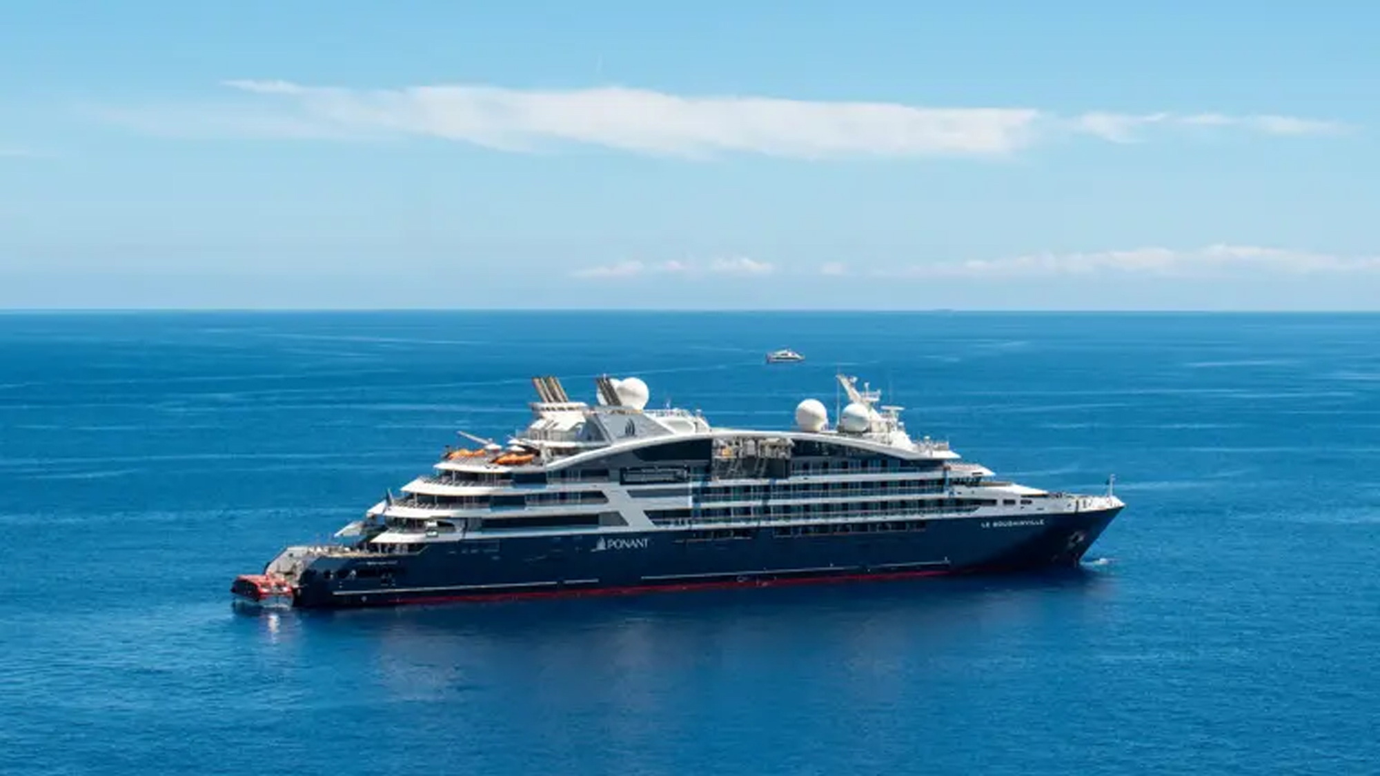 A sophisticated expedition cruise ship anchored in crystal-clear blue Mediterranean waters under a partly cloudy sky. The vessel features multiple decks with modern amenities, prominent communication and navigation equipment on the upper decks, and displays the characteristic livery of a luxury cruise operator with dark hull and white superstructure.