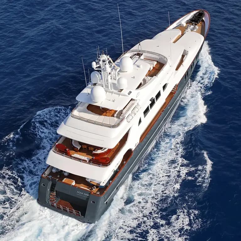 An aerial view of a multi-deck luxury expedition yacht navigating through deep blue ocean waters, featuring white and wood-tone hull design with visible wake trails. The vessel displays professional maritime equipment including communication antennas and appears to be underway in tropical or subtropical conditions.