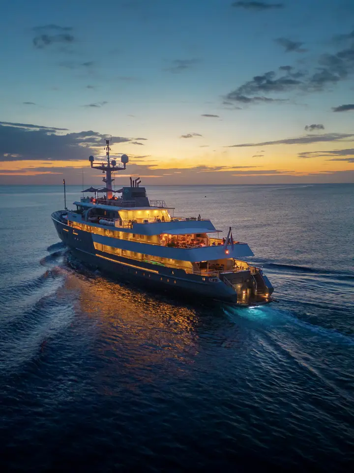 A sophisticated expedition yacht illuminated by warm golden lights during the blue hour at sunset, anchored in calm Arctic or northern waters. The vessel features multiple decks with illuminated windows and professional navigation equipment, representing luxury expedition cruising to remote polar destinations.