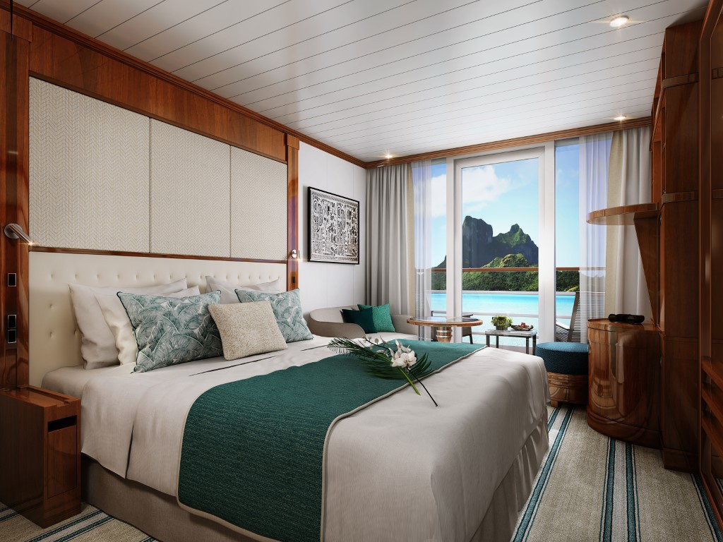 An elegantly appointed cruise ship cabin featuring a king-size bed with cream linens and teal accents, floor-to-ceiling windows offering panoramic views of turquoise waters and dramatic volcanic peaks, rich wood paneling, and contemporary furnishings that blend maritime style with tropical resort comfort.