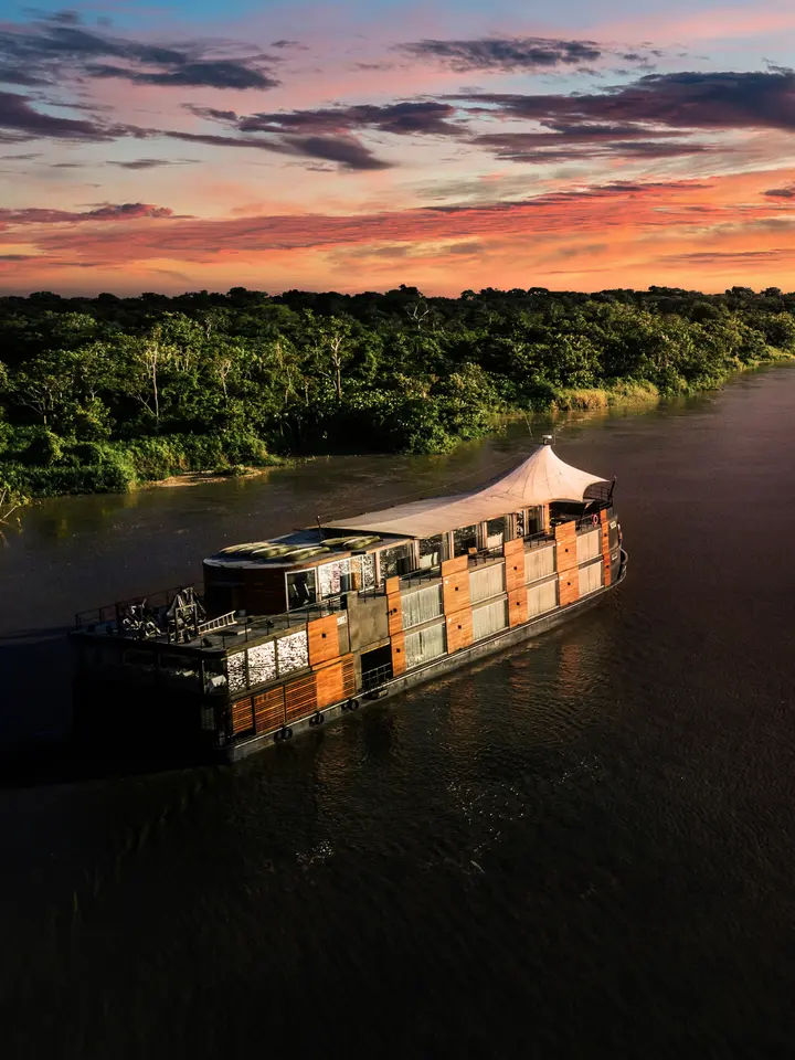 A distinctive wooden-hulled river cruise ship with orange and tan paneling navigates the Amazon River at golden hour, framed by dense rainforest canopy and a dramatic sunset sky filled with pink, orange, and purple clouds.