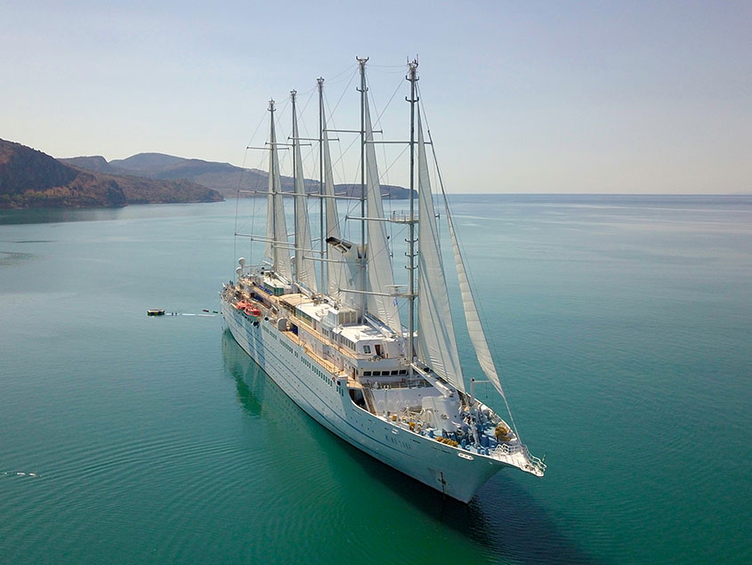 A stunning aerial view of a large multi-masted sailing cruise ship anchored in pristine turquoise waters near a mountainous coastal region. The vessel features multiple tall masts and white sails, with mountains visible in the background across calm, crystalline seas.