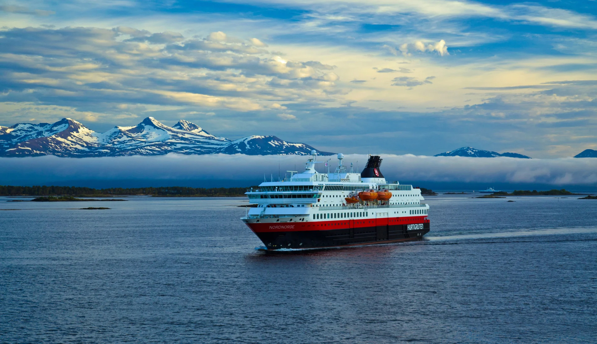 A striking image of the MS Nordnorge, a Norwegian coastal cruise ship, anchored in pristine Arctic waters with snow-capped mountains rising dramatically in the background. The vessel's distinctive red and black hull contrasts beautifully against the blue sea and sky, with scattered islands and towering peaks characteristic of Norway's northern regions.