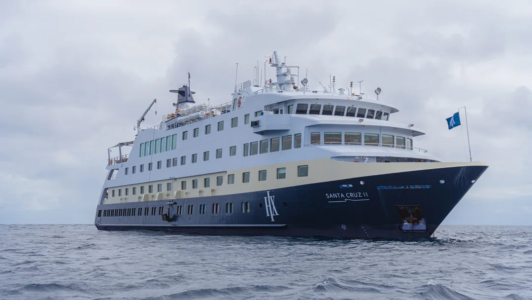 A modern expedition cruise ship with distinctive white and dark blue hull livery sailing on open ocean waters. The vessel features multiple passenger decks with numerous windows, communication equipment and antennas on top, and displays a flag on the starboard side under overcast skies.