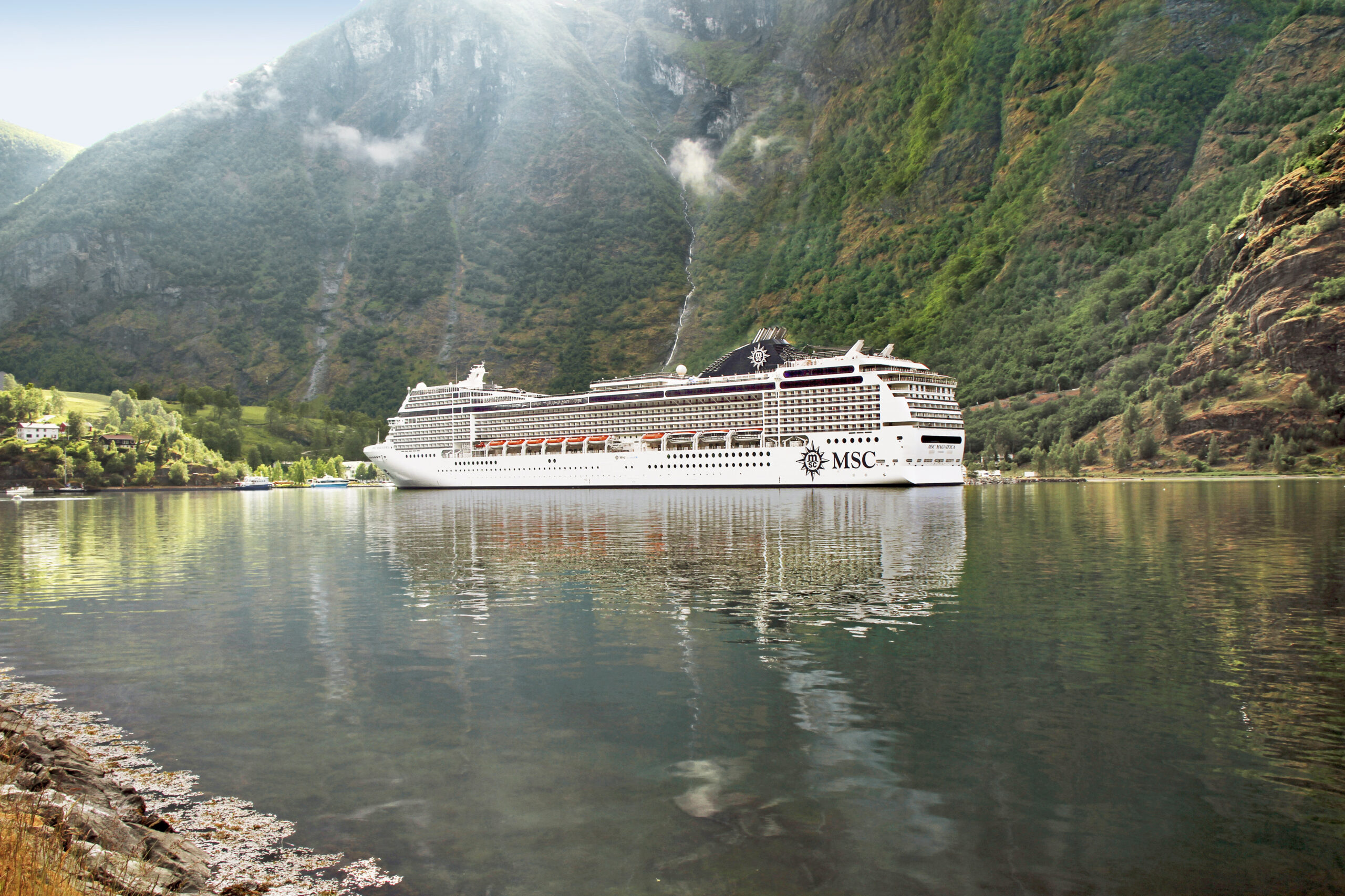 A large MSC cruise ship navigates through a dramatic Norwegian fjord surrounded by steep, forested mountain cliffs and cascading waterfalls. The vessel is anchored in calm waters with picturesque alpine scenery and misty peaks in the background.