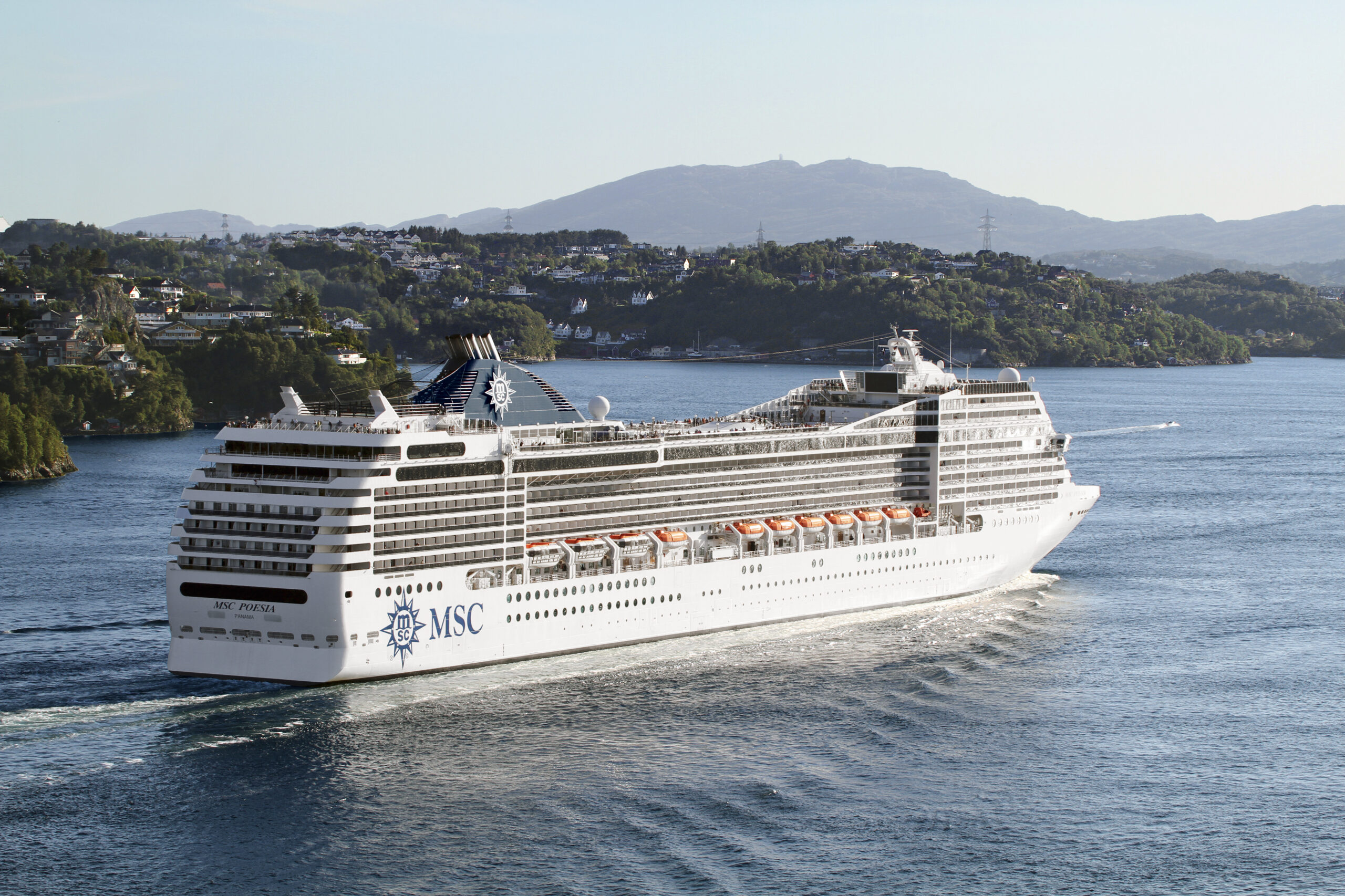 The MSC Poesia, a large modern cruise ship, sails through a picturesque Norwegian fjord surrounded by lush green mountains and waterfront settlements. The vessel's white hull and distinctive MSC branding are clearly visible as it navigates the serene blue waters with forested hillsides and residential areas dotting the landscape in the background.