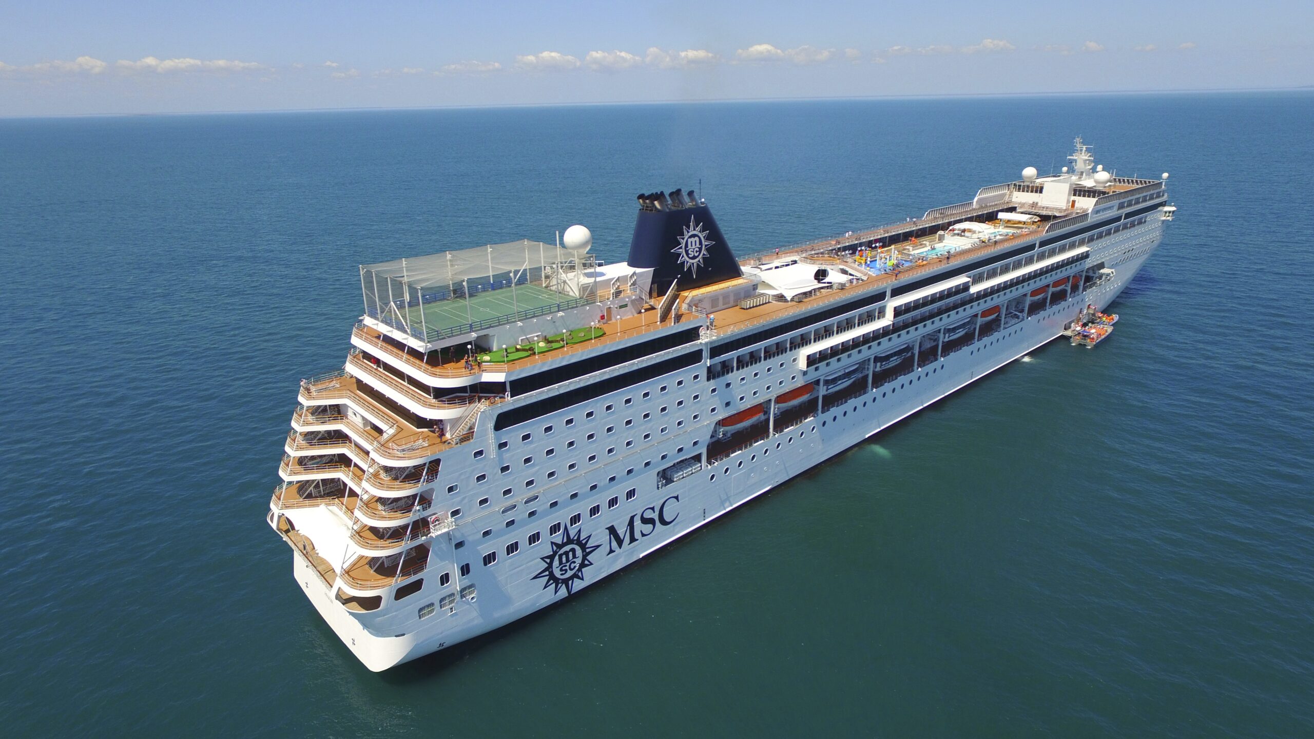 A modern cruise ship with distinctive MSC branding on the hull, photographed from an aerial perspective in calm blue-green waters. The vessel features multiple passenger decks, a sports court on the upper deck, and the characteristic MSC livery with a dark funnel bearing the ship's wheel logo.