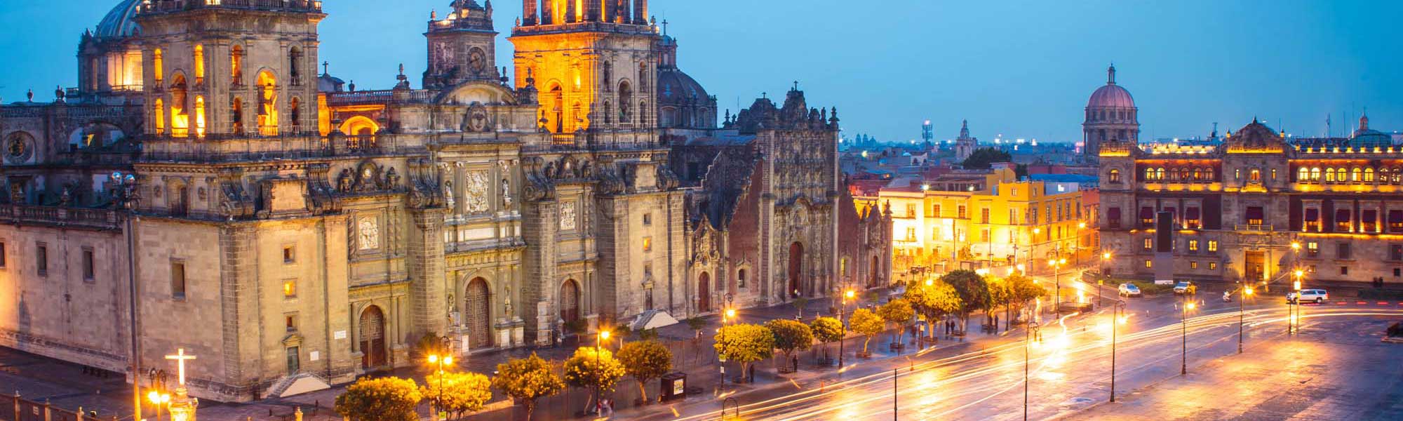 The magnificent Mexico City Metropolitan Cathedral illuminated against the twilight sky, showcasing stunning colonial architecture with golden lighting. The historic plaza features baroque stone facades, street lighting, and surrounding colonial buildings that reflect the rich architectural heritage of Mexico's capital city.