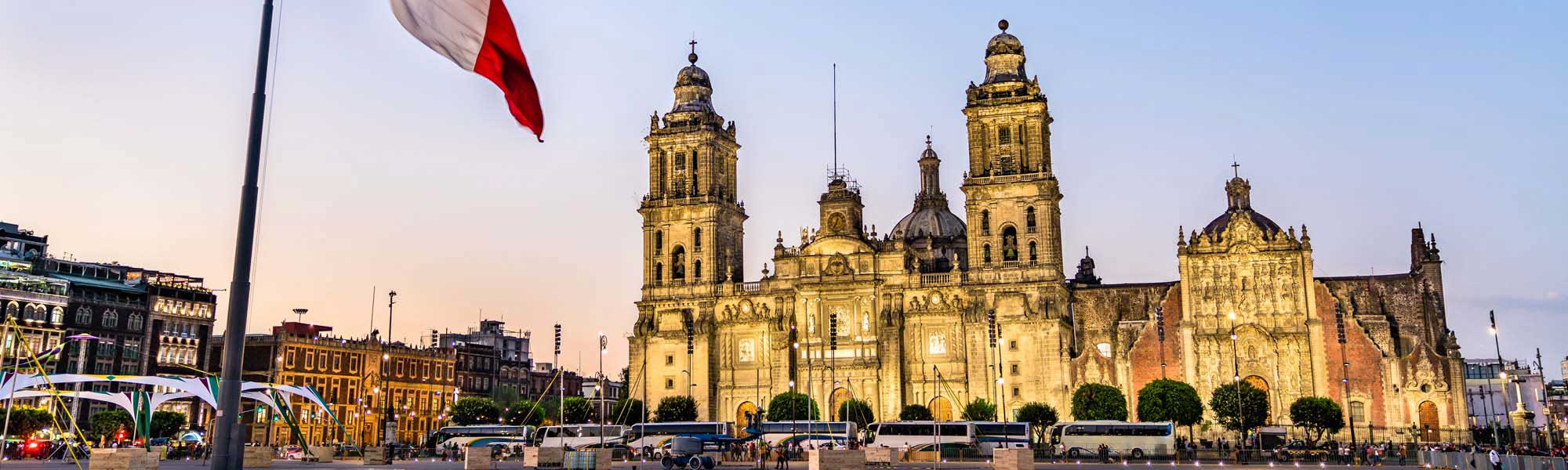 The iconic Mexico City Metropolitan Cathedral illuminated in golden light during twilight, featuring its distinctive Baroque architecture with twin towers and ornate facade. The plaza in front is bustling with visitors beneath the Mexican flag, showcasing one of North America's largest and oldest cathedrals.