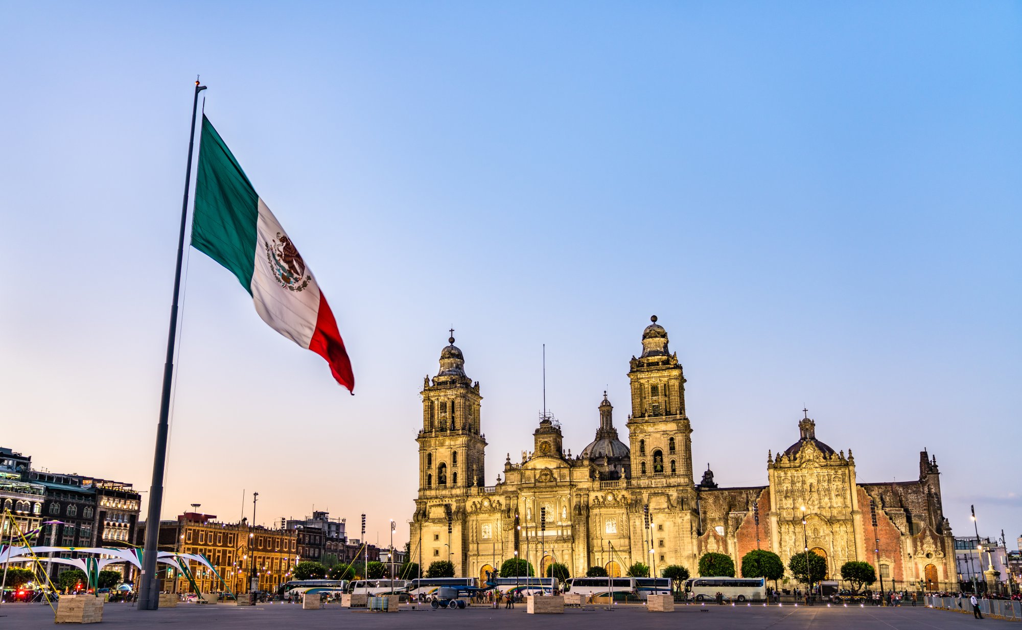 Mexico City Metropolitan Cathedral at Sunset The iconic Mexico City Metropolitan Cathedral illuminated during golden hour, with the Mexican flag prominently displayed in the foreground of the Zócalo plaza. The ornate Baroque architecture features twin bell towers and a distinctive dome, representing one of the largest and oldest cathedrals in the Americas.