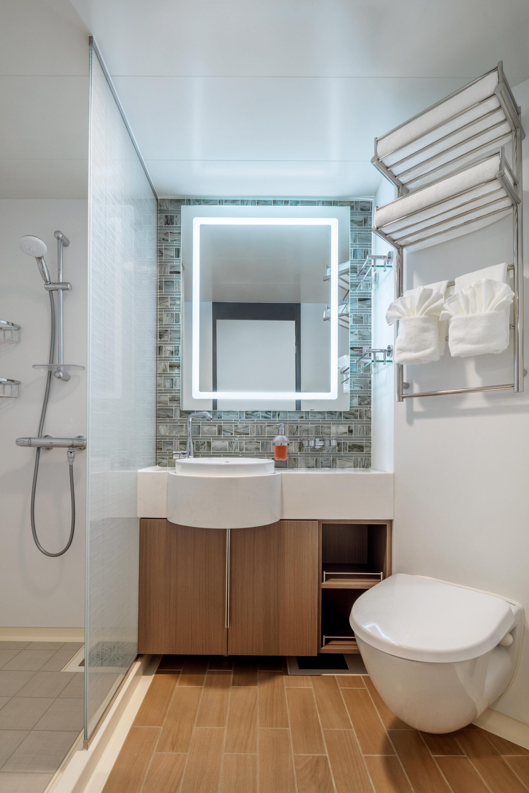 Modern Cruise Ship Balcony Stateroom Bathroom A contemporary cruise ship bathroom featuring a curved white ceramic sink, illuminated square mirror with LED lighting, and wood-grain vanity cabinetry. The space showcases modern amenities including a wall-mounted towel warmer, rainfall shower system, and decorative mosaic tile backsplash in neutral tones, exemplifying upscale cruise accommodation design.