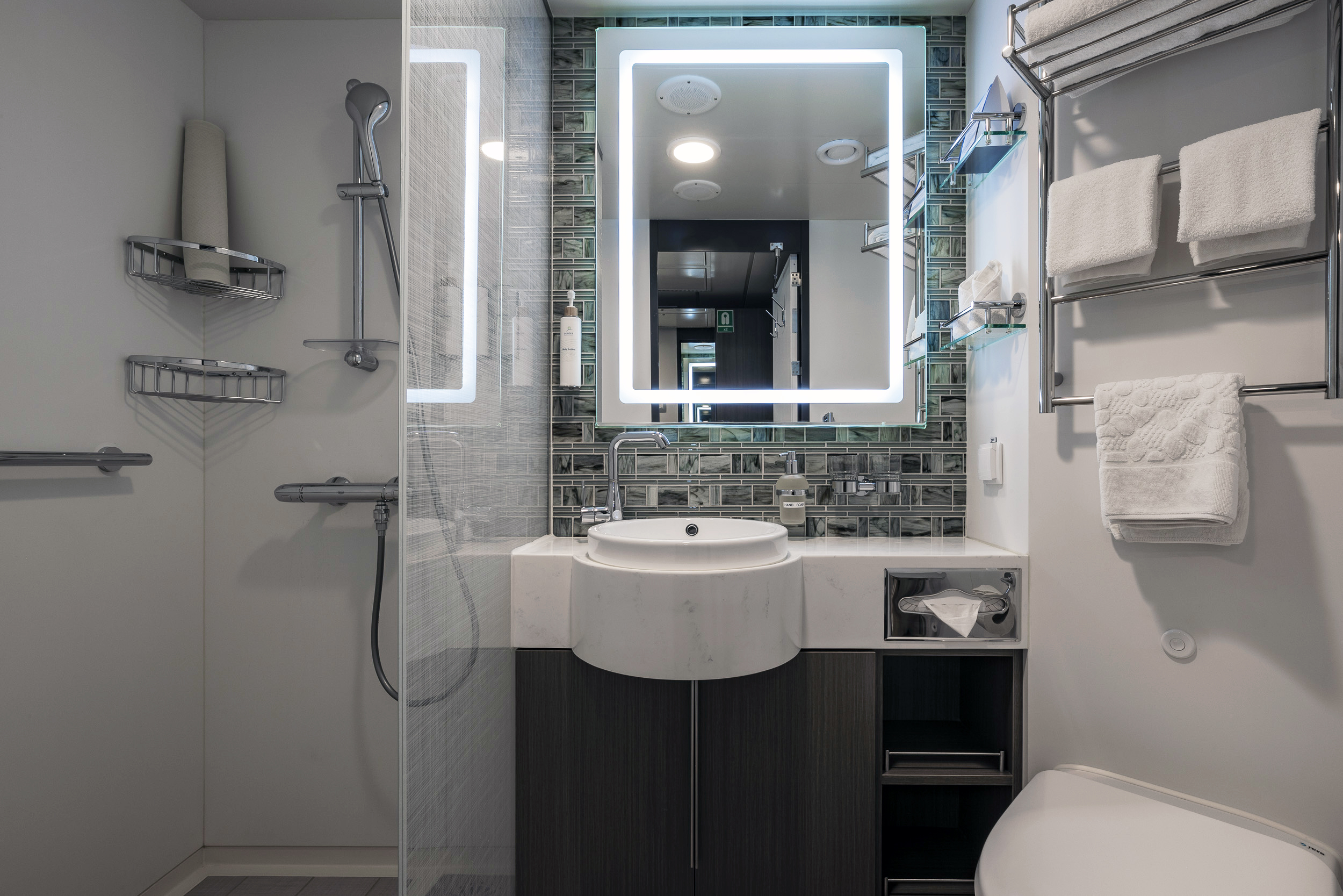 Modern Cruise Ship Bathroom with LED Mirror A contemporary bathroom aboard a cruise ship featuring a white curved sink, illuminated LED mirror, mosaic tile backsplash, and stainless steel towel racks. The space showcases modern amenities typical of a superior stateroom, with organized storage and neutral color palette.