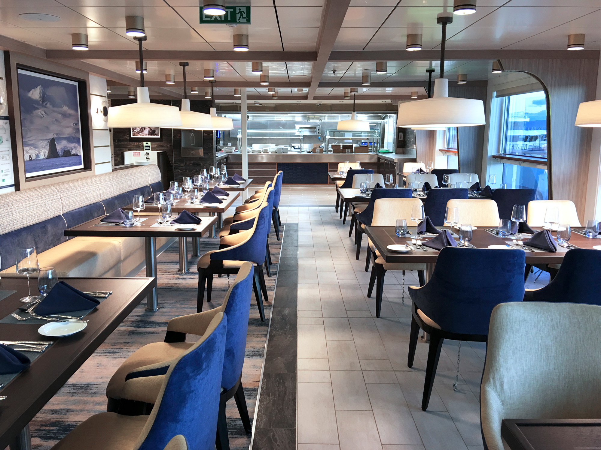 Modern Cruise Ship Dining Room with Ocean Views An elegant fine dining restaurant aboard a cruise ship, featuring contemporary design with blue velvet chairs, dark wood tables, pendant lighting, and floor-to-ceiling windows overlooking the ocean. The space showcases modern Scandinavian-influenced interior design with a professional open kitchen visible in the background.