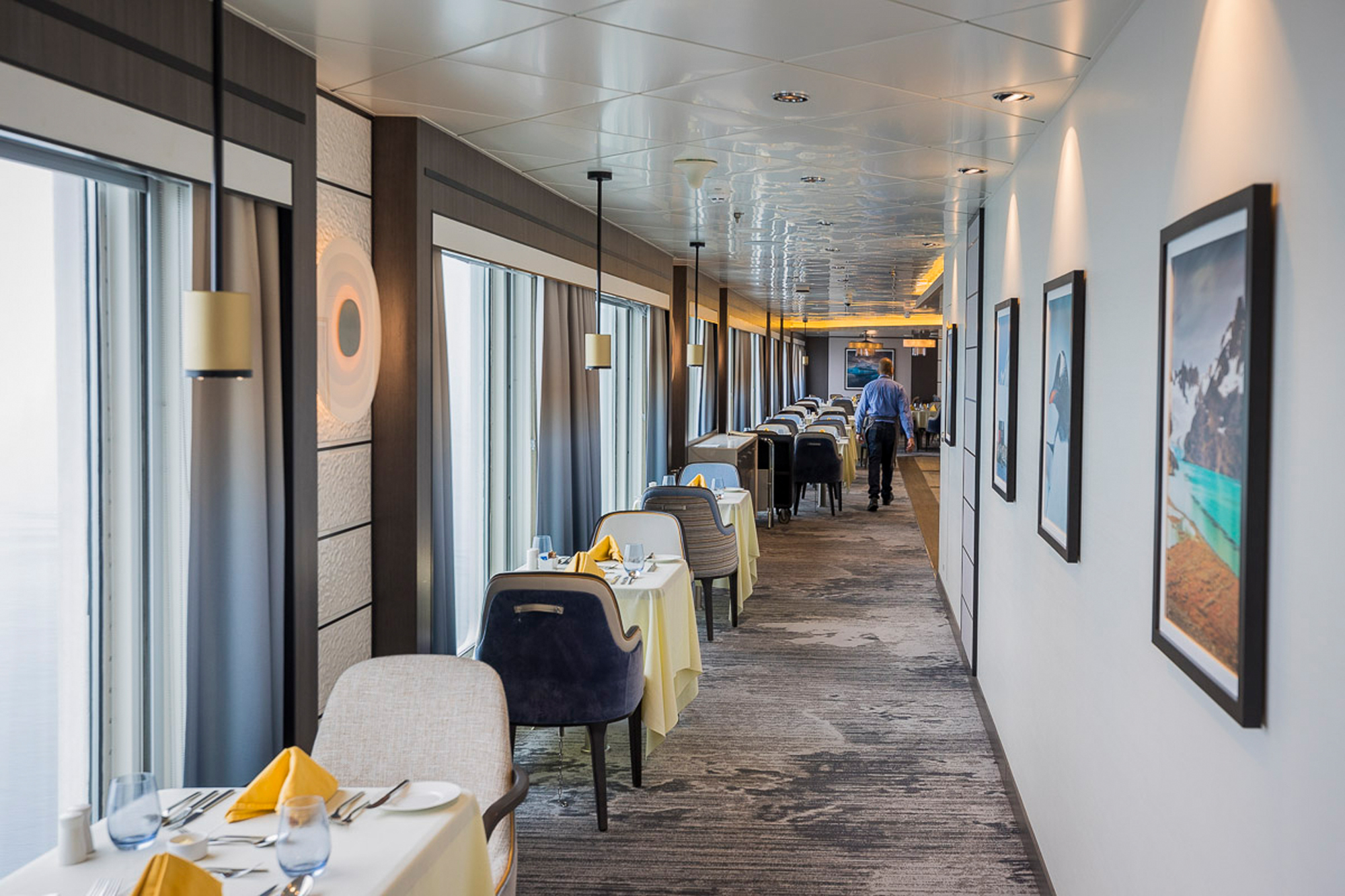 Modern Cruise Ship Dining Room with Ocean Views An elegant dining room aboard a modern cruise ship featuring floor-to-ceiling windows along the corridor, contemporary furniture with dark and cream upholstery, warm ambient lighting, and framed scenic artwork on the walls. The space showcases sophisticated interior design with a long perspective view down the dining corridor lined with tables set for service.