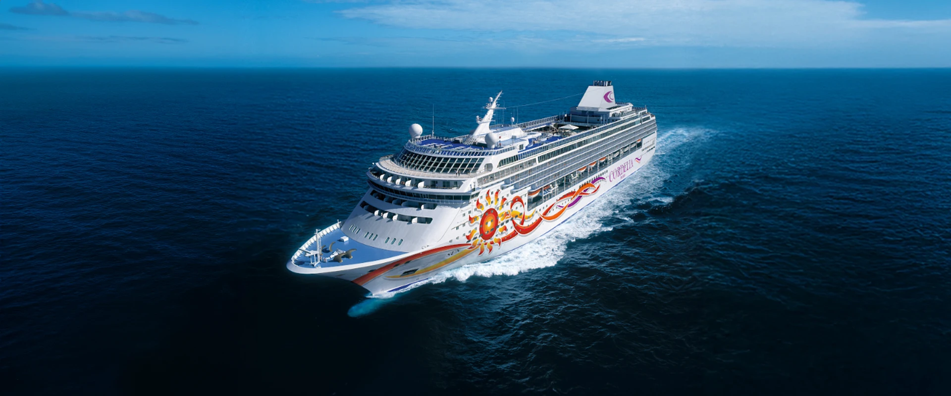 Modern Cruise Ship at Sea A large modern cruise ship with distinctive white and blue hull livery featuring colorful sun-themed artwork on its side, photographed from an aerial perspective while sailing through open ocean waters under clear blue skies.