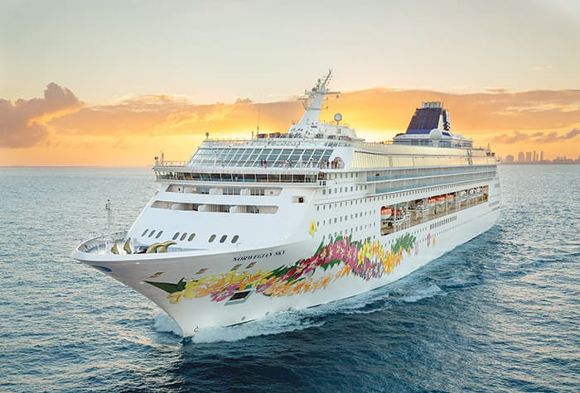 Modern Cruise Ship at Sunset A large, modern cruise ship with distinctive colorful floral artwork on its hull is photographed at sea during golden hour, with a vibrant sunset sky in the background and a coastal city visible on the horizon. The ship features multiple decks, contemporary design elements, and appears to be in tropical or subtropical waters.