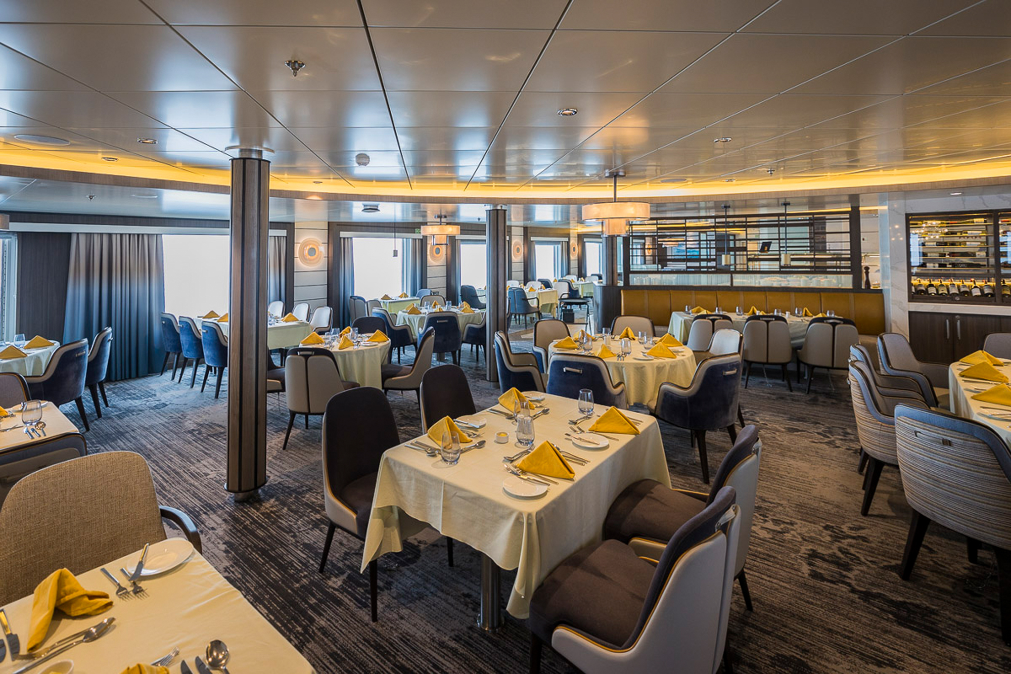 Modern Dining Room on Expedition Cruise Ship An elegant and spacious dining room aboard an expedition cruise vessel, featuring round tables with cream-colored linens and yellow napkins, contemporary beige and black chairs, and a sophisticated interior with metallic ceiling panels and ambient lighting. The venue showcases modern design elements including support columns, large windows, and a refined ambiance suitable for fine dining at sea.