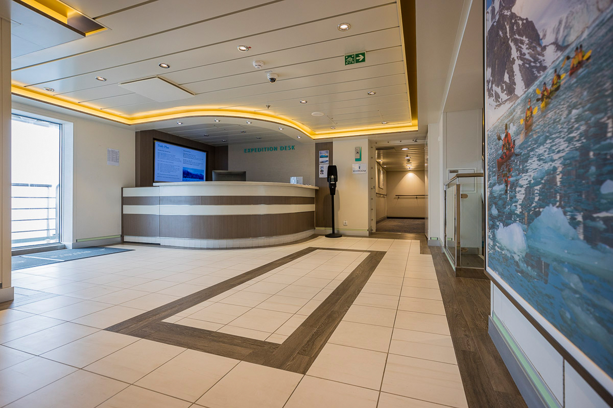 Modern Expedition Desk Lobby Interior A contemporary expedition desk in a modern cruise ship or hotel lobby, featuring a curved reception counter with brown and white paneling, warm golden ceiling lighting, and large-format expedition photography displayed on the walls. The space exemplifies modern hospitality design with clean lines, professional signage, and welcoming architectural elements.