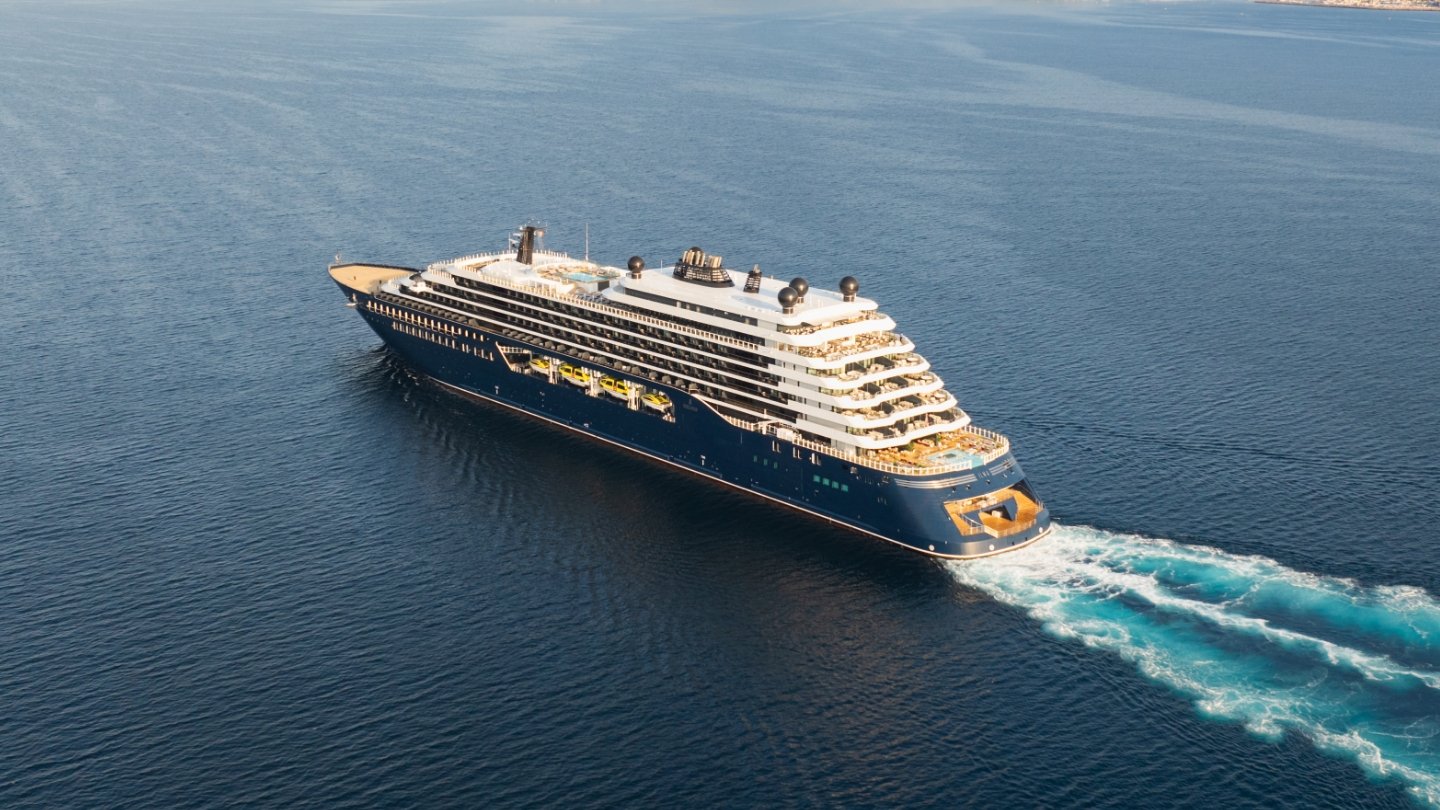 A large modern cruise ship with distinctive dark blue and white hull features multiple decks with golden-hued upper structures, photographed sailing through calm blue waters with white wake visible behind it.