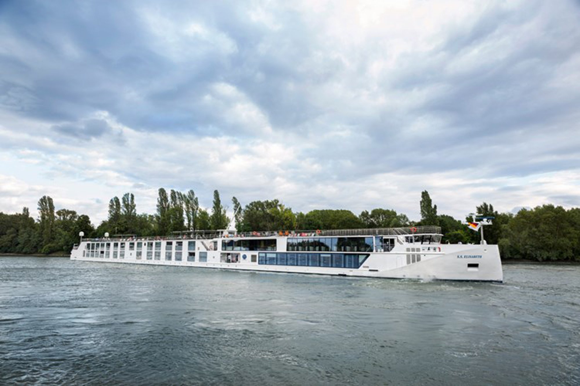 A contemporary all-white river cruise ship navigates a calm waterway lined with lush green vegetation and poplar trees. The vessel features multiple passenger decks with extensive panoramic windows, modern amenities, and a distinctive streamlined design typical of European river cruising.