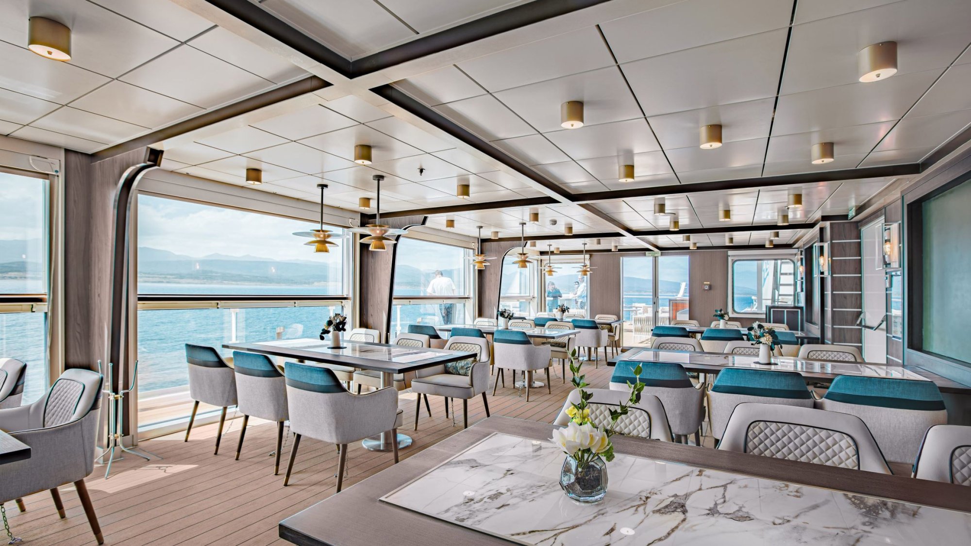 An elegant specialty dining venue aboard a cruise ship featuring floor-to-ceiling ocean views, contemporary furniture with teal and gray upholstery, marble-topped tables, and a sophisticated coffered ceiling with brass accents. The space showcases panoramic sea vistas with coastal mountains visible in the distance.