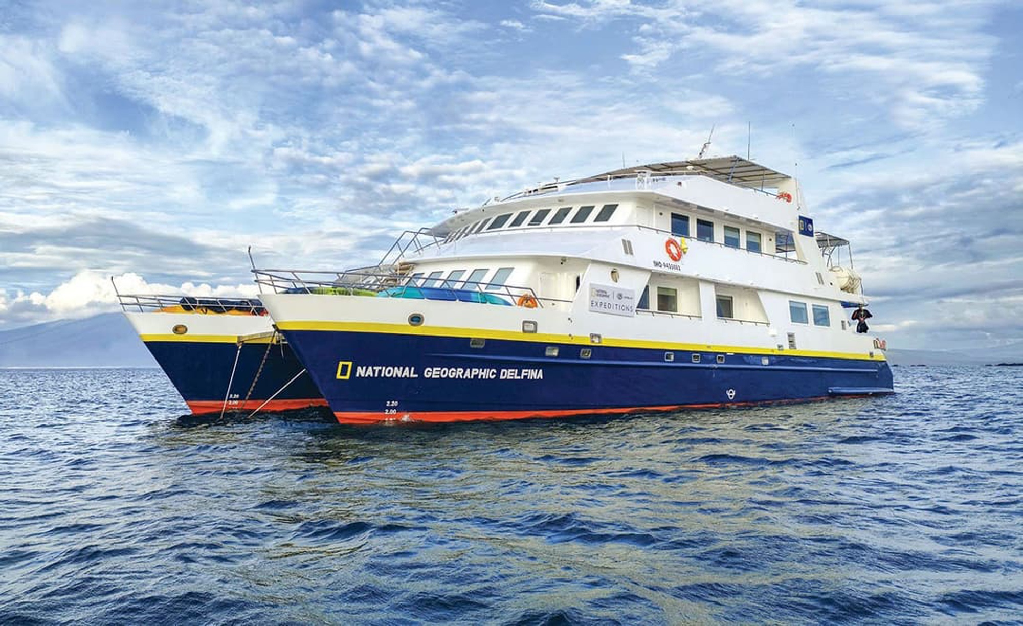 National Geographic Delfina Expedition Cruise Ship The National Geographic Delfina, an expedition cruise vessel, is anchored in clear tropical waters with a distinctive white and navy blue hull featuring a yellow stripe. The ship is equipped with multiple decks, a tender boat alongside, and visible National Geographic branding, designed for luxury expedition travel to remote destinations like the Galápagos Islands.
