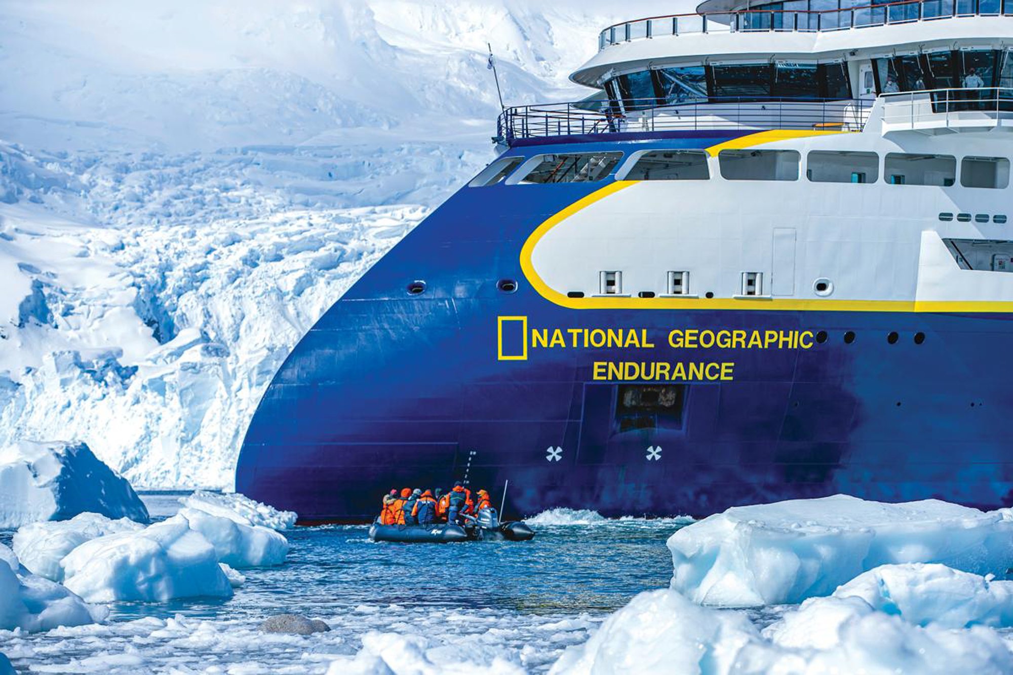 National Geographic Endurance in Antarctic Waters The National Geographic Endurance expedition cruise ship navigates through icy Antarctic waters, with a zodiac full of passengers in orange life jackets exploring the pristine iceberg-filled landscape. Snow-covered mountains and massive ice formations frame this polar exploration scene.