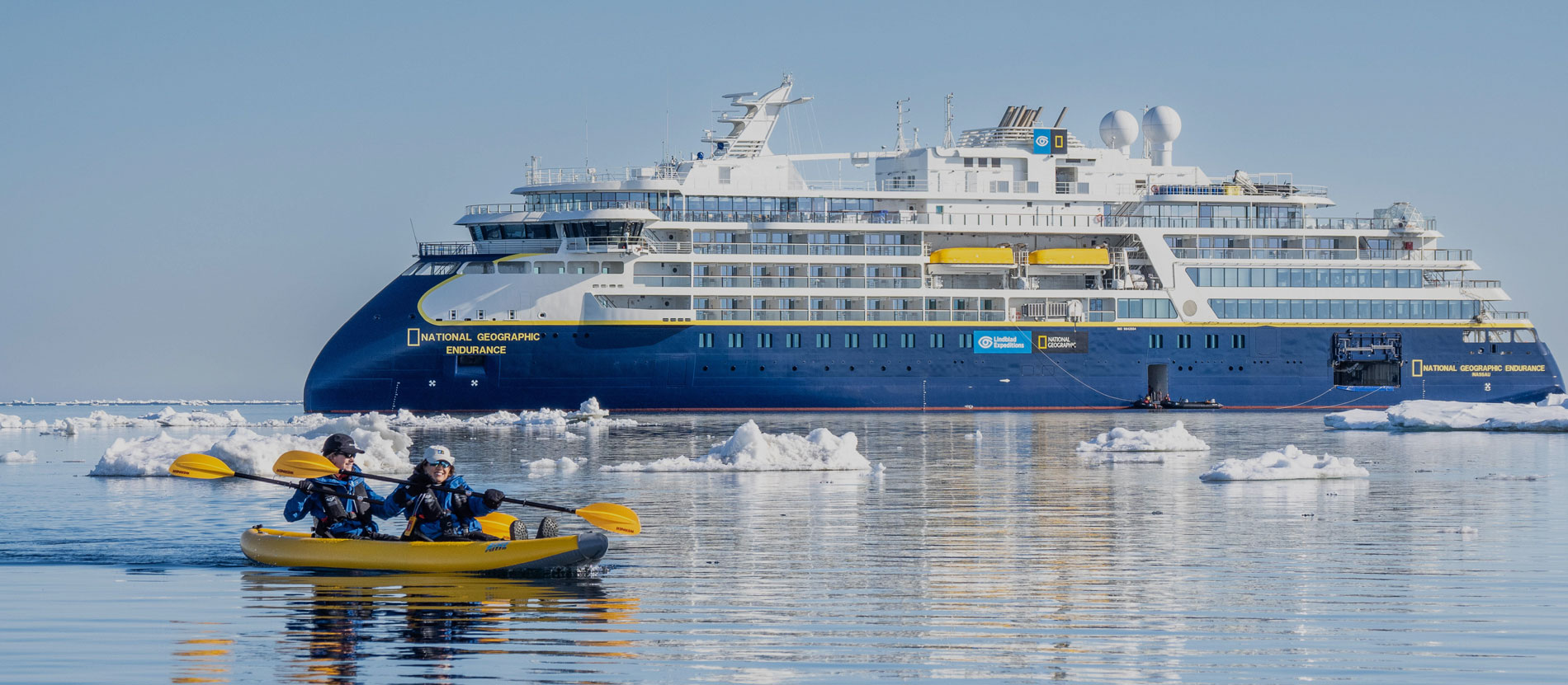 National Geographic Endurance in Arctic Ice The National Geographic Endurance expedition cruise ship navigates through icy Arctic waters while two kayakers in a yellow kayak explore the pristine polar landscape dotted with white ice chunks. This image showcases the contrast between the modern expedition vessel and adventurous water activities in one of the world's most remote destinations.