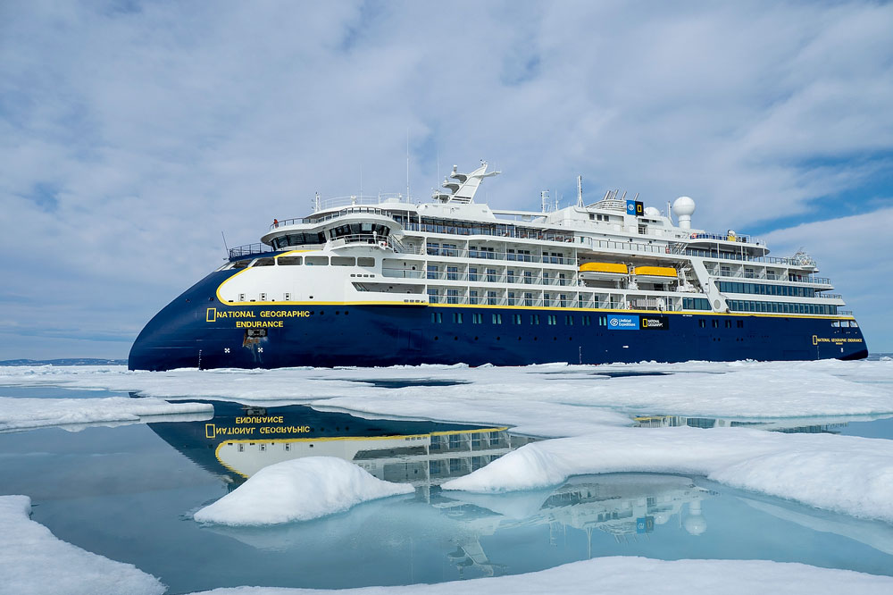 National Geographic Endurance in Arctic Ice The National Geographic Endurance expedition cruise ship navigates through Arctic pack ice, surrounded by pristine white ice floes and clear blue meltwater channels. The vessel's distinctive dark blue and yellow hull is reflected in the calm water, showcasing modern expedition cruising in extreme polar environments.