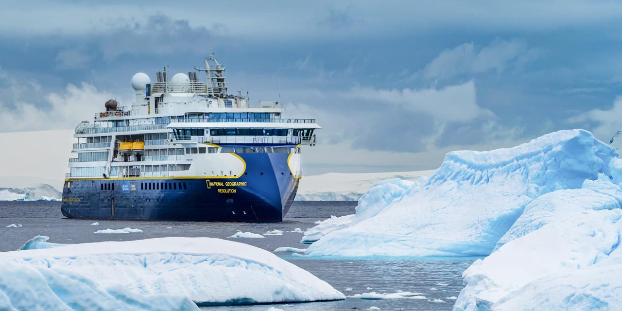 National Geographic Resolution in Antarctic Waters The National Geographic Resolution expedition cruise ship navigates through icy Antarctic waters surrounded by massive blue icebergs in the Gerlache Strait. The vessel, identifiable by its distinctive blue and yellow hull and National Geographic branding, is framed by towering glacial ice formations under a partly cloudy sky.