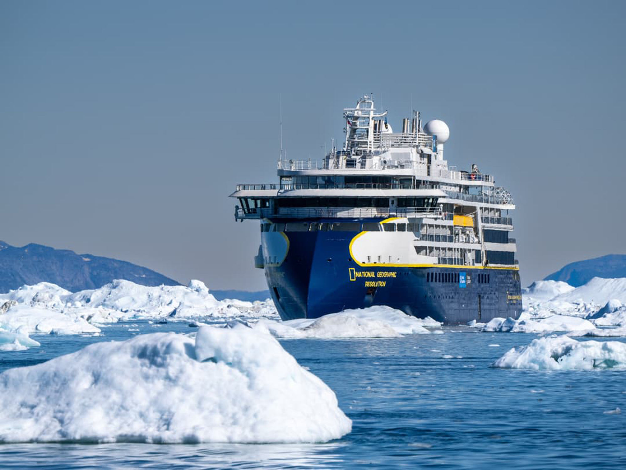 National Geographic Resolution in Arctic Ice The National Geographic Resolution expedition cruise ship navigates through icy Arctic waters surrounded by large icebergs and glacial ice floes. The modern expedition vessel, identifiable by its distinctive blue hull and National Geographic branding, is positioned among the floating ice in what appears to be a Greenland or Arctic fjord setting.