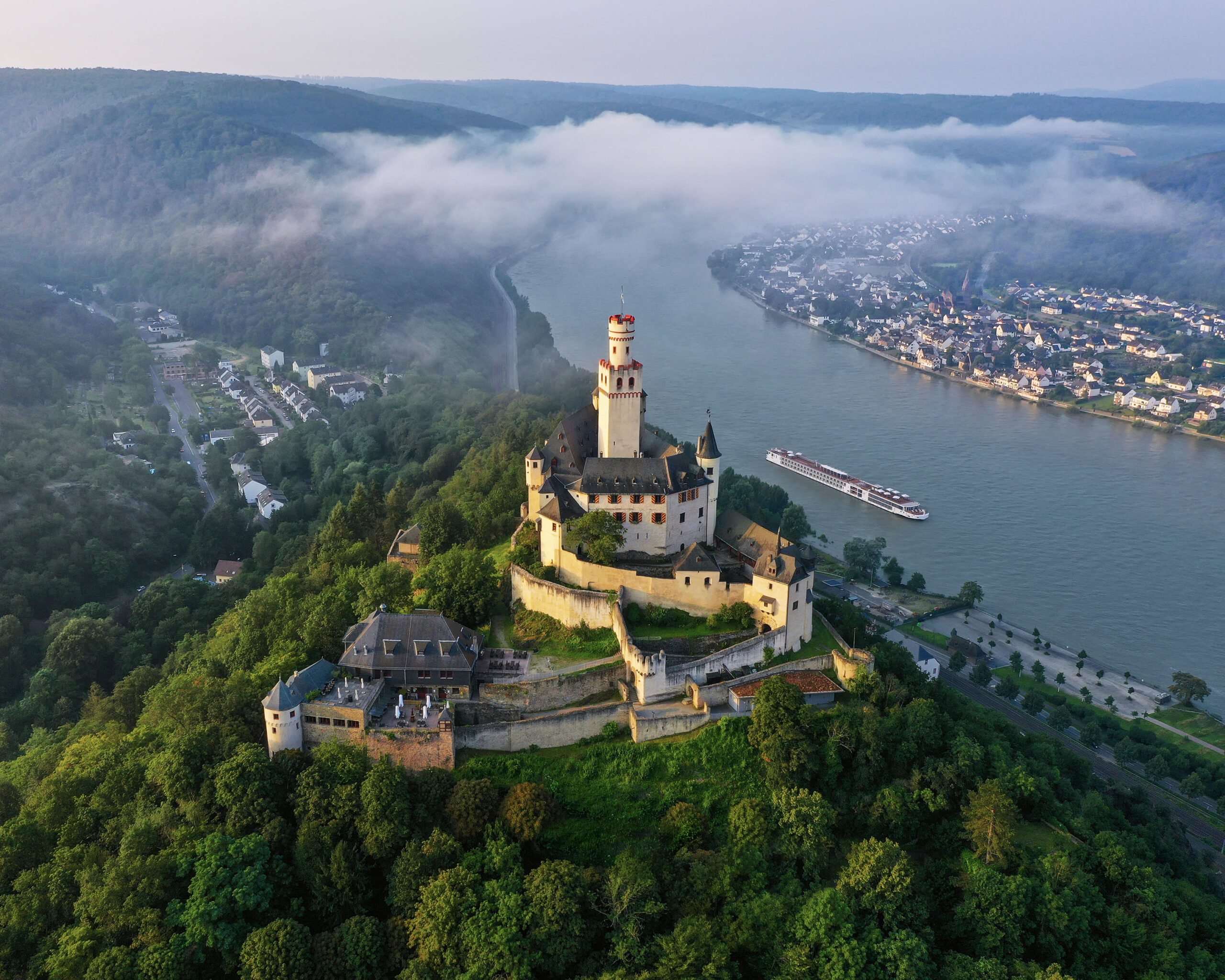 An aerial view of the iconic Neuschwanstein Castle perched on a hilltop above the Rhine River in Germany, with a river cruise ship navigating the misty valley below. The castle's distinctive yellow limestone walls and towers rise majestically from dense forests, while morning fog blankets the picturesque riverside towns and vineyards along the river bend.