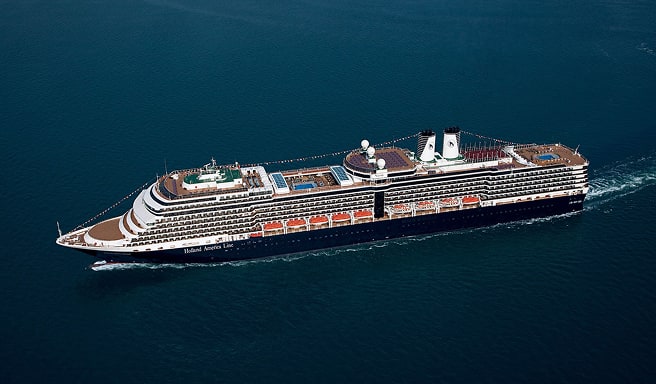 Aerial photograph of the Nieuw Amsterdam, a large modern cruise ship, sailing through calm turquoise waters. The ship displays distinctive Holland America Line livery with multiple decks, a white superstructure, and red hull accents visible along its side.