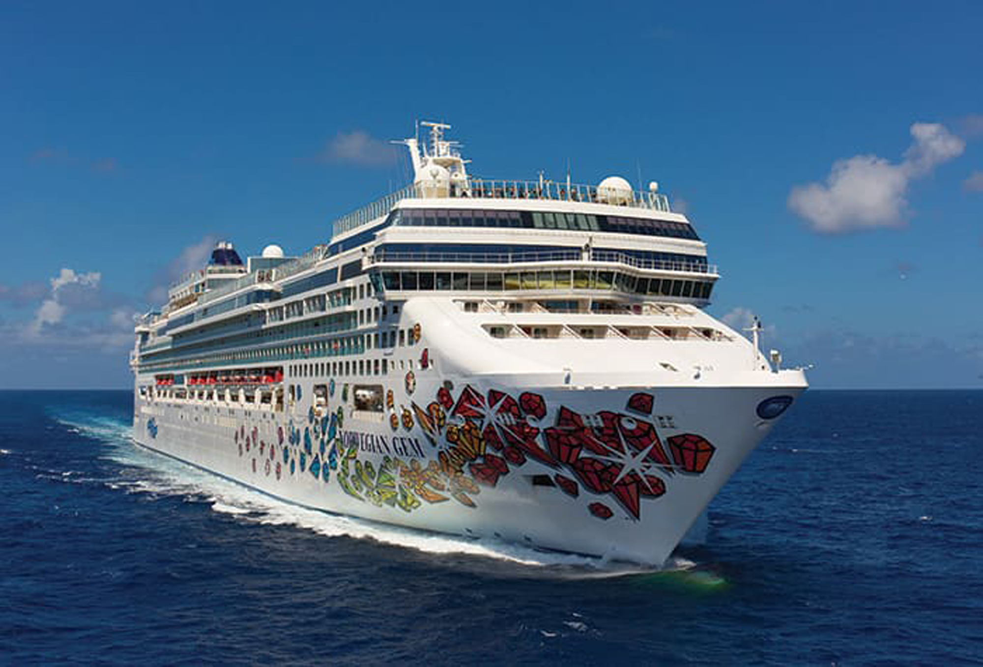 Norwegian Gem Cruise Ship at Sea A large modern cruise ship with distinctive red and gold artistic hull graphics sailing on calm blue ocean waters under clear skies. The multi-deck vessel features a white superstructure with multiple levels of balconies and amenities visible along its profile.