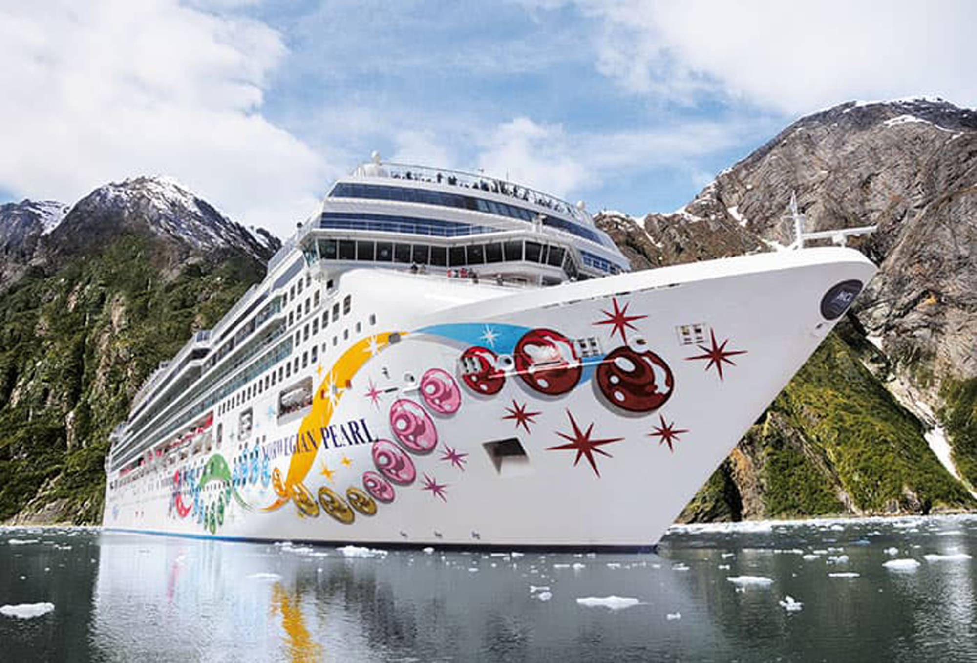Norwegian Pearl in Glacier Bay, Alaska A large cruise ship navigates through a pristine Alaskan glacier bay, flanked by towering snow-capped mountains and dense forest. The vessel displays colorful artwork on its hull featuring festive designs in red, yellow, and pink tones, with the ship's name 'PEARL' visible on the bow.
