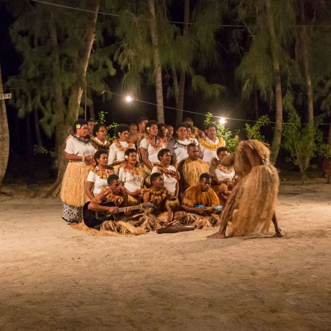 A vibrant nighttime cultural gathering on a sandy beach featuring a large group of performers in traditional Pacific Islander attire, including grass skirts and flower leis, arranged in a circle under string lights and towering palm trees. The scene captures an authentic ceremonial or entertainment experience typical of island excursions offered by cruise lines visiting the South Pacific.