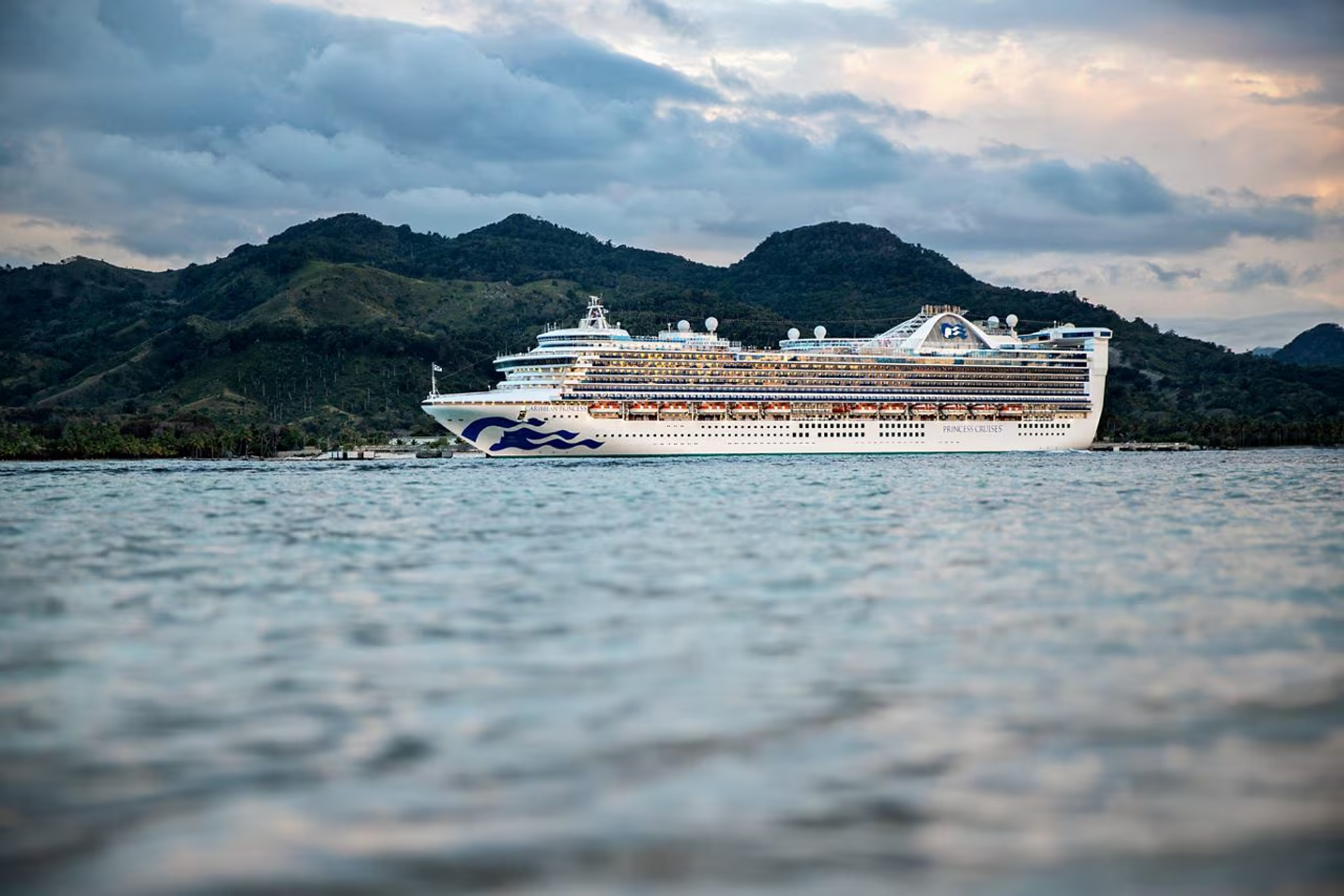 Princess Cruises Ship at Tropical Port A large modern cruise ship with distinctive Princess Cruises livery anchored in calm turquoise waters, with lush green tropical mountains in the background under a dramatic cloudy sky.