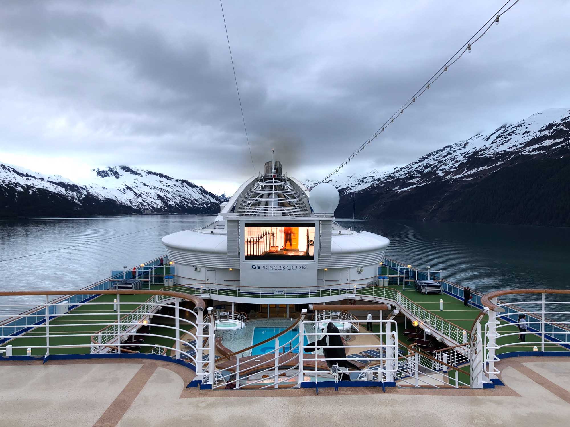 Princess Cruises Ship in Glacier Bay Alaska A Princess Cruises vessel navigates through a snow-capped fjord in Alaska, with the ship's distinctive dome structure and warm interior lighting visible against the dramatic glacial landscape. Snow-covered mountains frame both sides of the waterway, creating a striking contrast between the modern cruise ship and pristine wilderness.