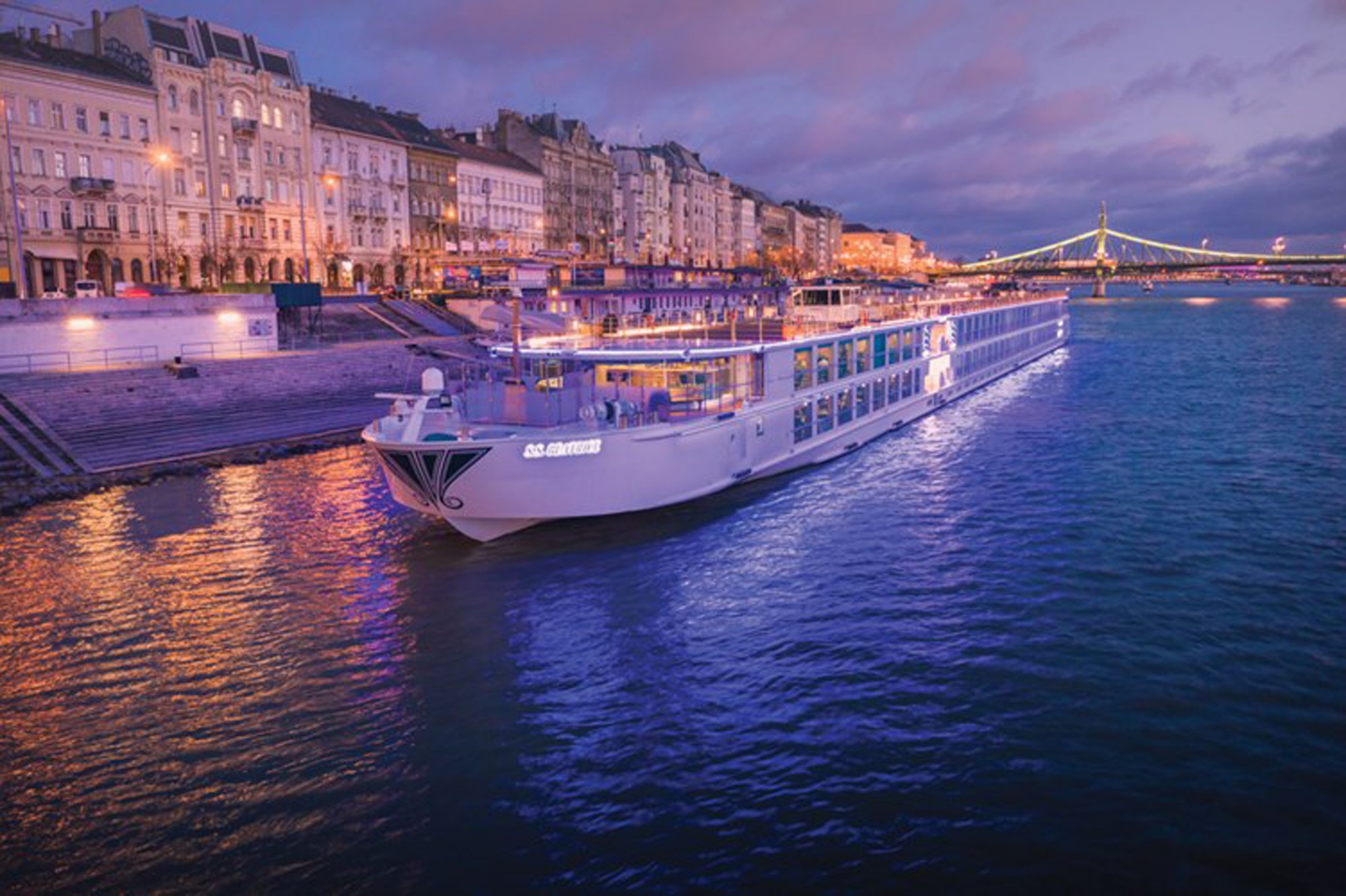 A modern river cruise ship is docked along the Danube River in Budapest during twilight hours, with illuminated classical European architecture lining the waterfront and the iconic Liberty Bridge visible in the background across the water.
