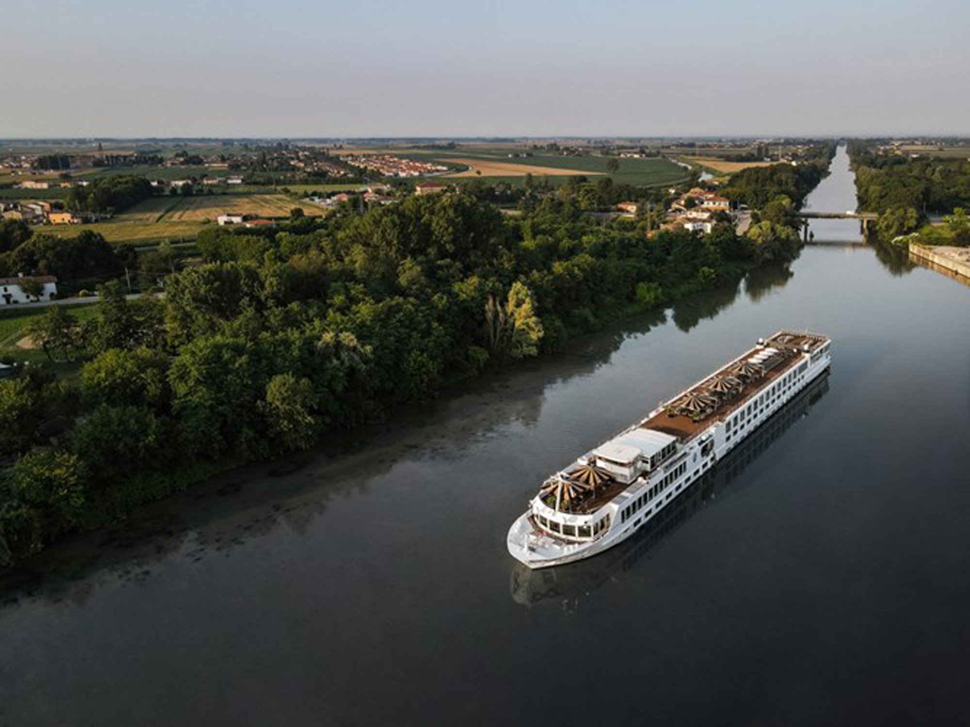 A modern river cruise ship navigates a picturesque European inland waterway, with lush green forests lining the banks and a charming countryside landscape visible in the distance, featuring agricultural fields, small villages, and a bridge crossing the calm water.