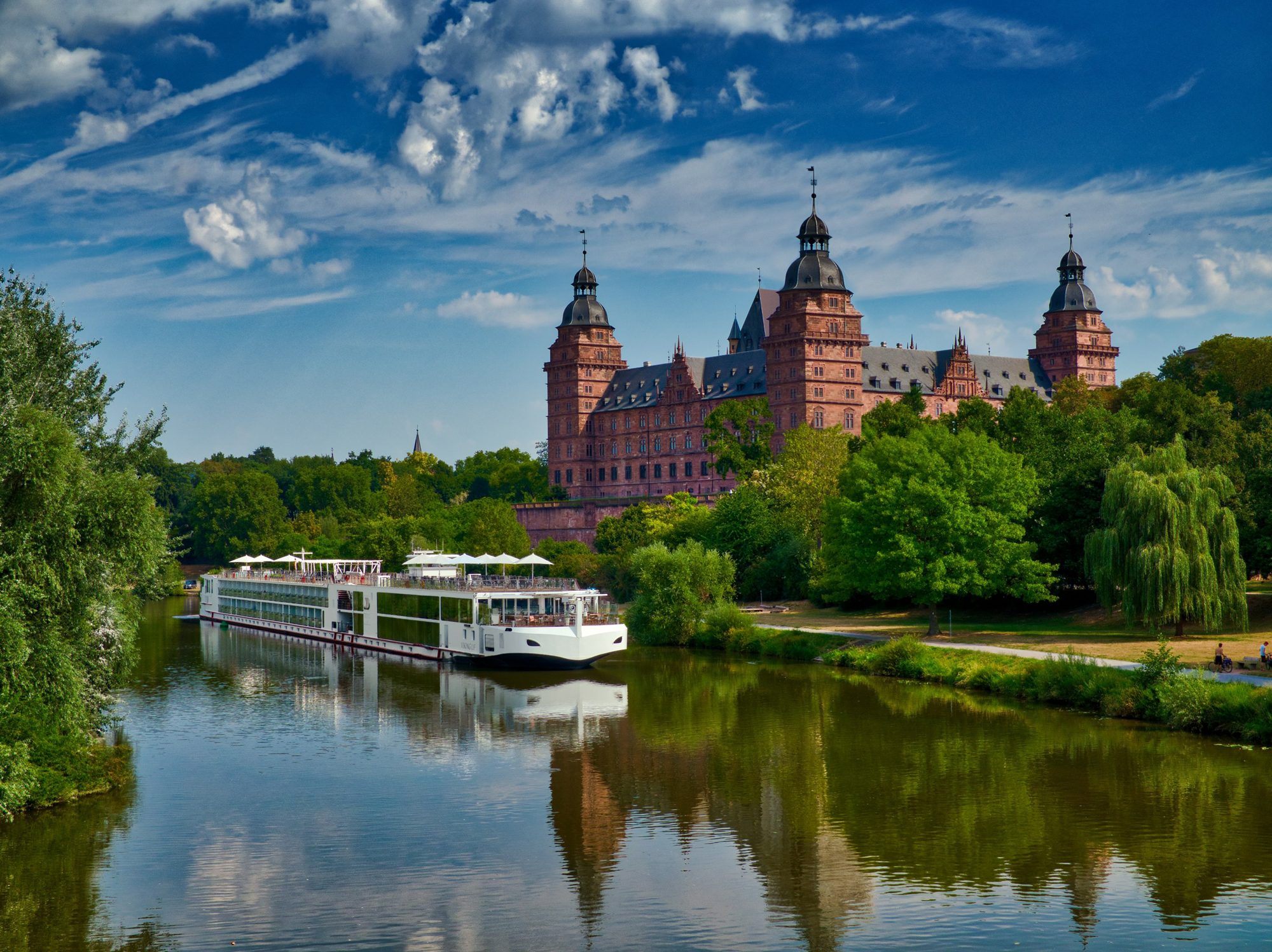 A modern river cruise ship navigates the Main River in Aschaffenburg, Germany, with the stunning red-sandstone Aschaffenburg Palace rising majestically in the background. The elegant vessel is framed by lush green riverbanks under a beautiful blue sky with white clouds, showcasing the scenic beauty of German river cruising.