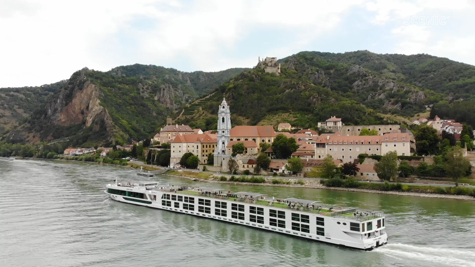 A modern river cruise ship navigates the Danube River past the picturesque Austrian town of Dürnstein, featuring a prominent white church steeple, terracotta-roofed buildings, and dramatic limestone cliffs dotted with vineyards and a castle ruin perched on the hilltop.