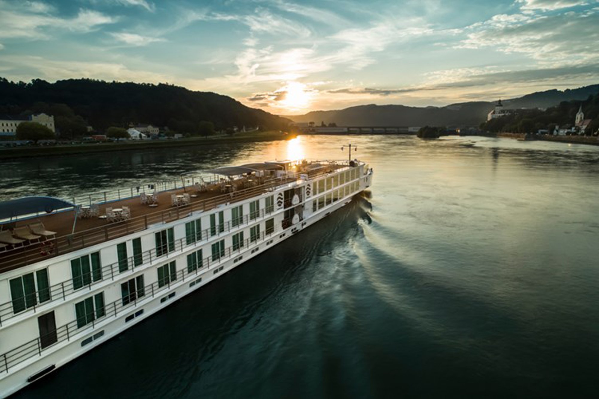 A modern river cruise ship with white and green exterior design navigates a picturesque European river valley at golden hour, with forested hills, a bridge, and charming riverside villages visible in the background under a beautiful sunset sky.