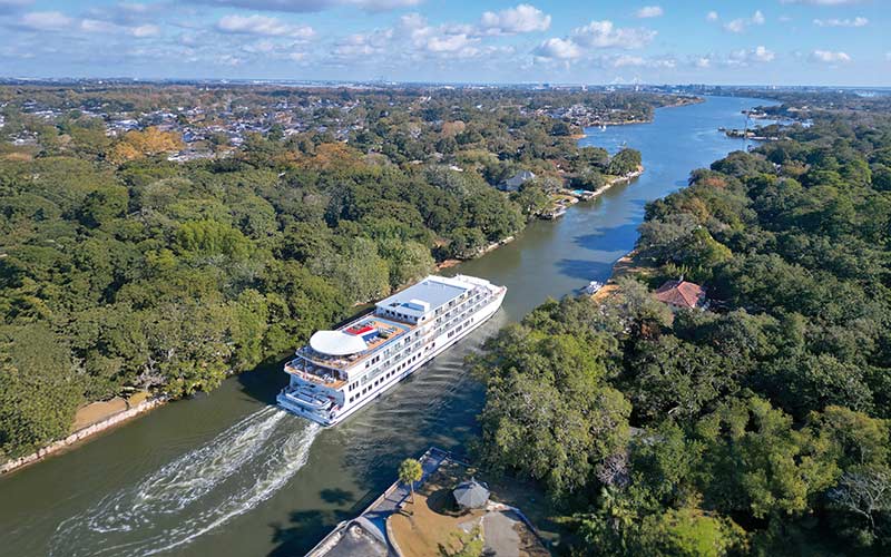 River Cruise Ship in Tropical Waterway An aerial view of a multi-deck river cruise ship navigating a narrow waterway surrounded by lush green forests and residential areas. The vessel is traveling through what appears to be a tropical or subtropical river system with clear blue water and dense vegetation on both banks.