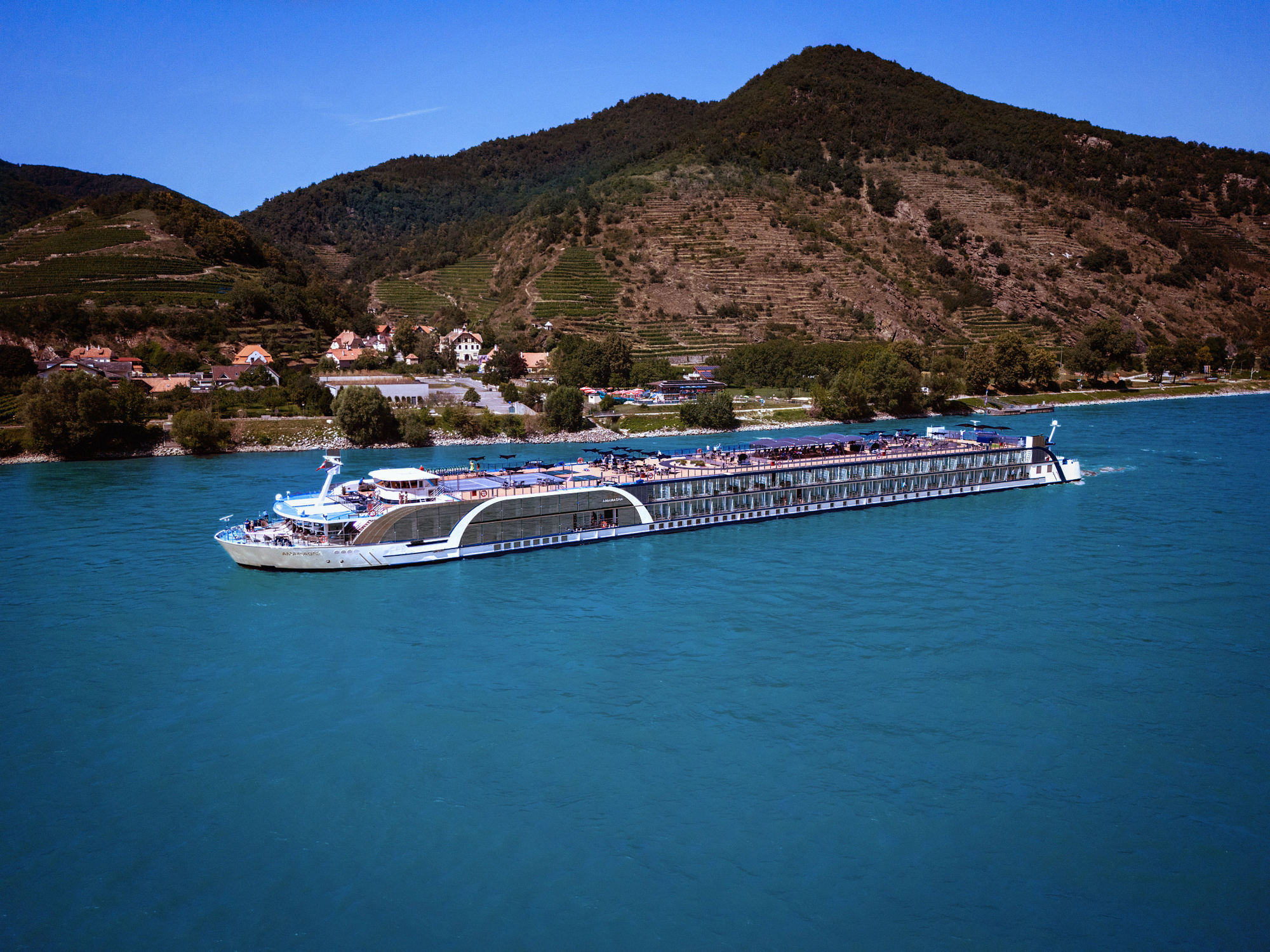 A modern river cruise ship navigates the turquoise waters of the Danube River, passing through a picturesque Alpine valley with terraced vineyards on steep hillsides and a charming riverside village with traditional architecture nestled against forested mountains under clear blue skies.