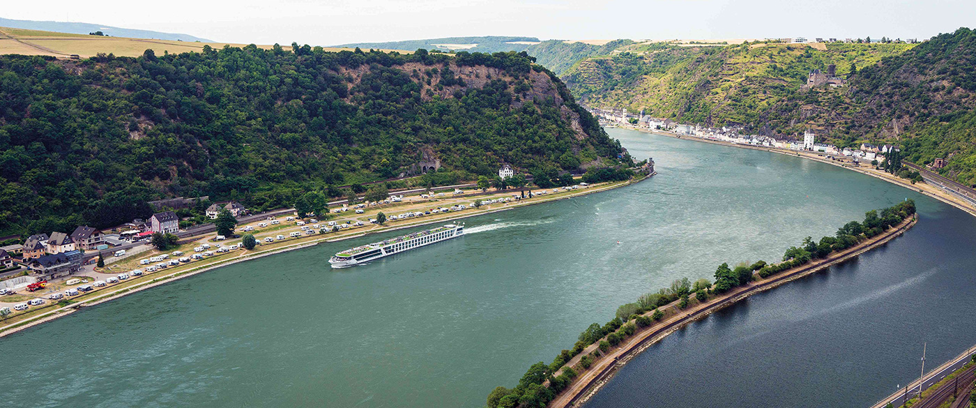 River Cruise Through Rhine River Gorge An aerial view of a modern river cruise ship navigating through the dramatic Rhine River Gorge in Germany, with steep forested cliffs on both sides, charming riverside towns nestled below, and lush green vegetation covering the hillsides.
