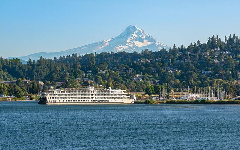 Riverboat Cruising with Mount Hood A classic riverboat cruise ship navigates the Columbia River with snow-capped Mount Hood rising dramatically in the background, surrounded by dense Pacific Northwest forest and waterfront homes nestled along the shoreline.