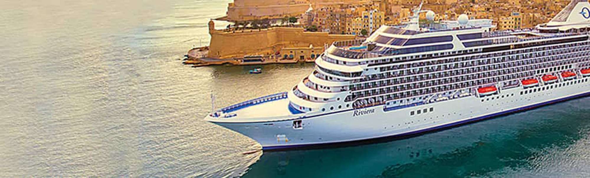 A modern cruise ship with blue and white livery is moored at a Mediterranean port, with the historic fortified harbor and golden limestone buildings of Valletta, Malta visible in the background. The ship features multiple passenger decks with distinctive curved balconies and a sleek bow design.