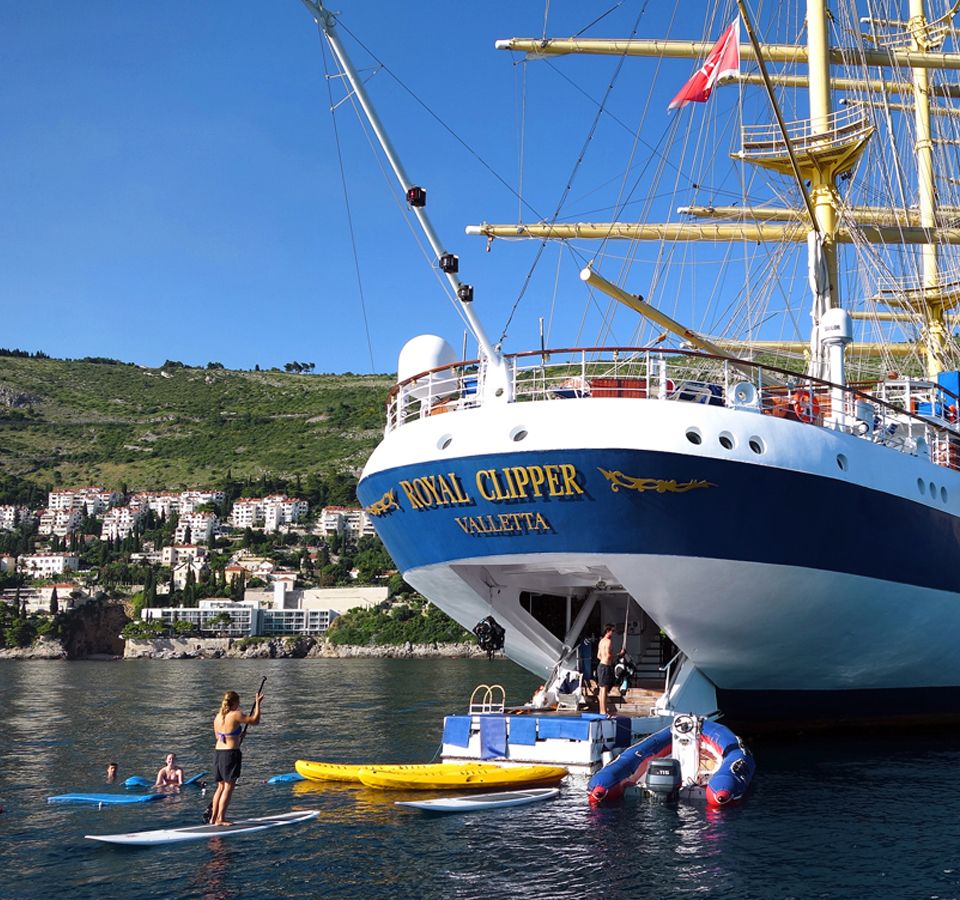 The tall ship Royal Clipper Valletta is anchored in a Mediterranean harbor with passengers enjoying watersports activities including paddleboarding and jet skis in the clear blue water. The vessel's distinctive square-rigged sails and masts are visible against a hillside dotted with Mediterranean architecture.